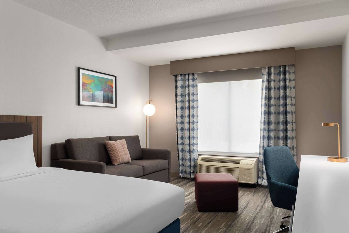 A hotel room with a bed, a sofa, a desk, a chair, a lamp, a picture on the wall, and a window with curtains.