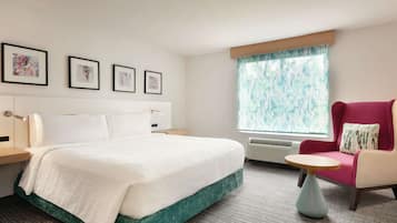 A hotel room with a bed, a chair, a small table, and a window with curtains.