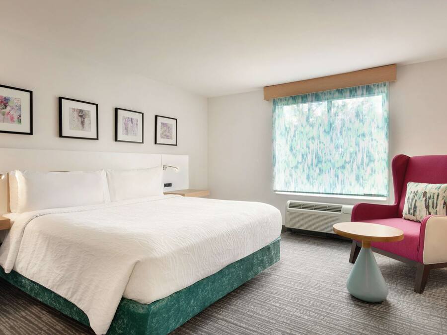 A hotel room with a bed, a chair, a small table, and a window with curtains.
