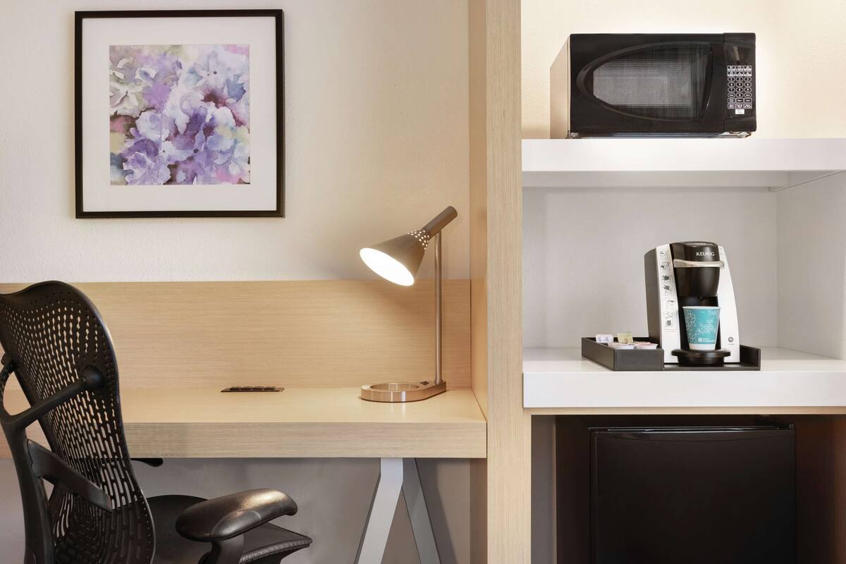 A hotel room with a desk, a chair, a framed floral artwork, a microwave, and a coffee machine.