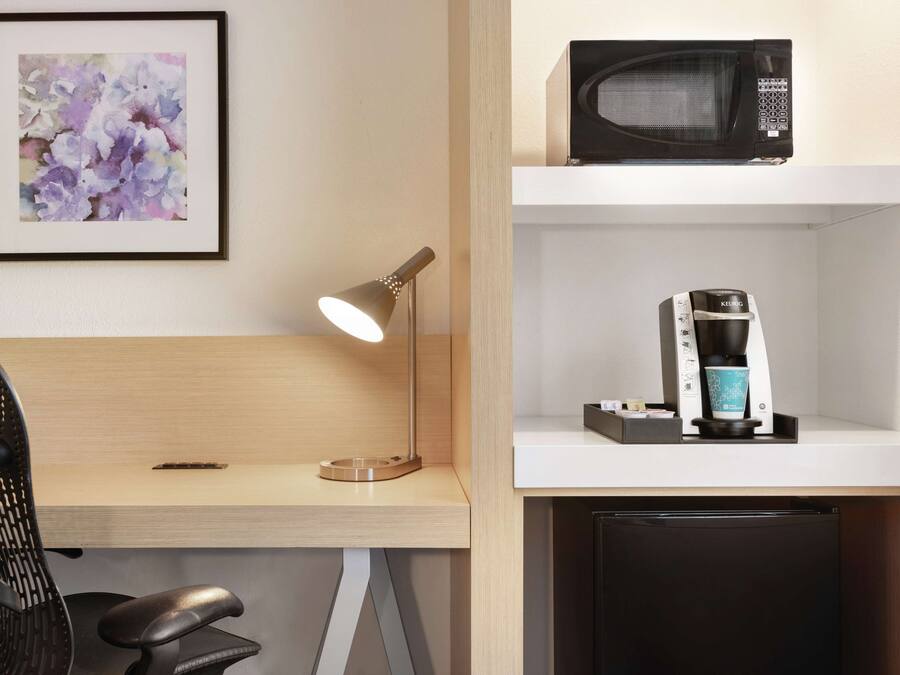 A hotel room with a desk, a chair, a framed floral artwork, a microwave, and a coffee machine.