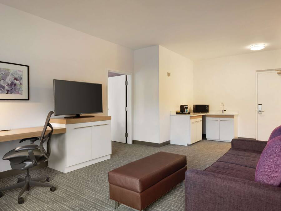 Une chambre d’hôtel moderne équipée d’un téléviseur à écran plat, d’un bureau, d’un pouf marron, d’un canapé violet et d’une kitchenette.