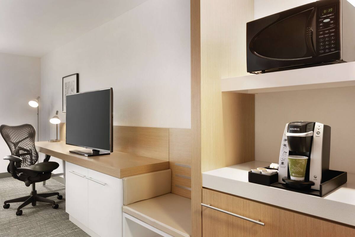 A modern hotel room with a desk, a computer, a chair, a microwave, and a coffee maker.