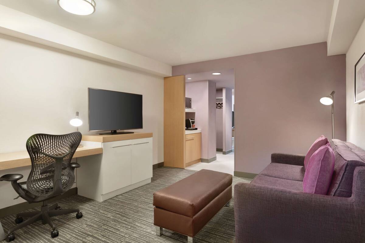 A modern hotel room with a desk, office chair, sofa, and a television.