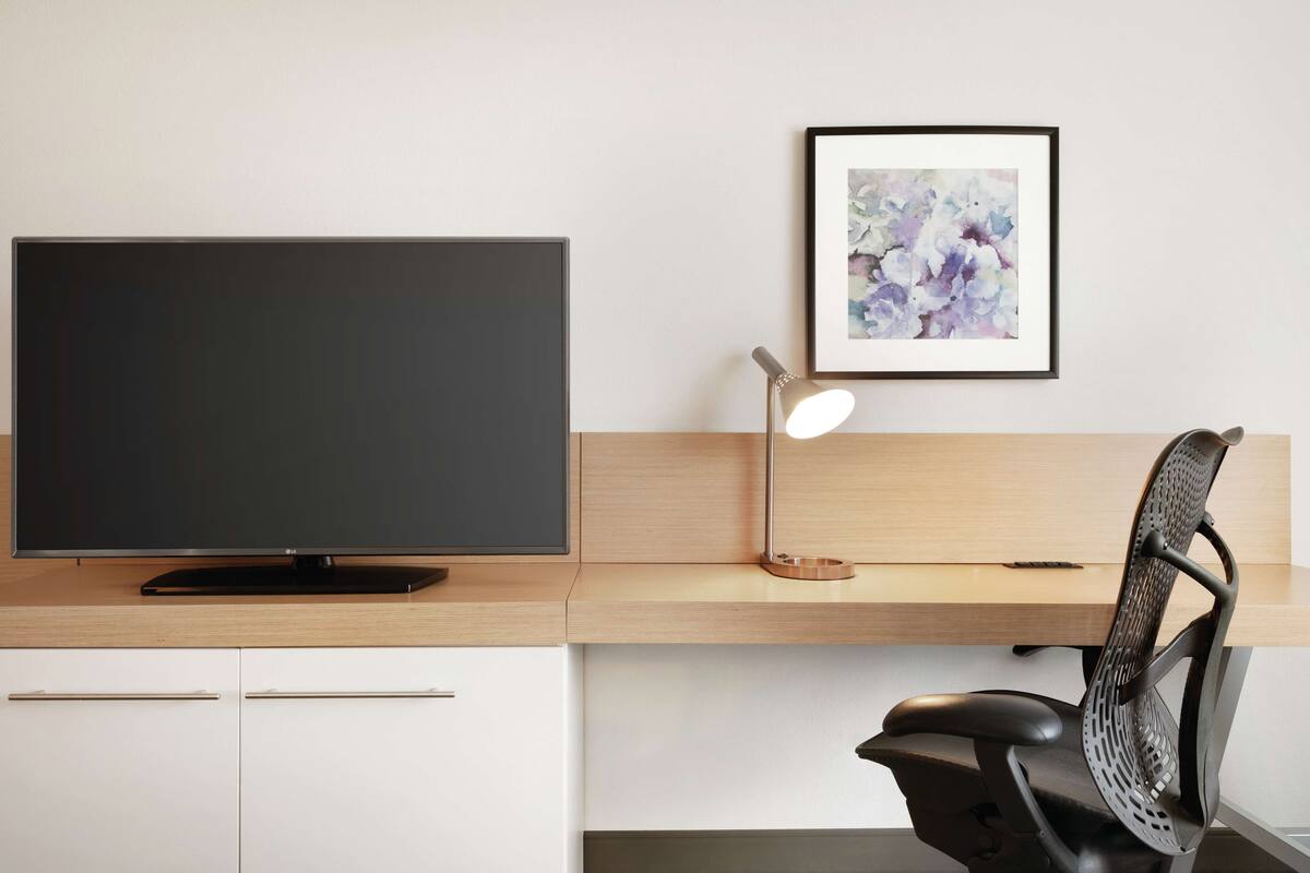 A modern office desk with a flat-screen TV, a desk lamp, and a framed abstract painting on the wall.