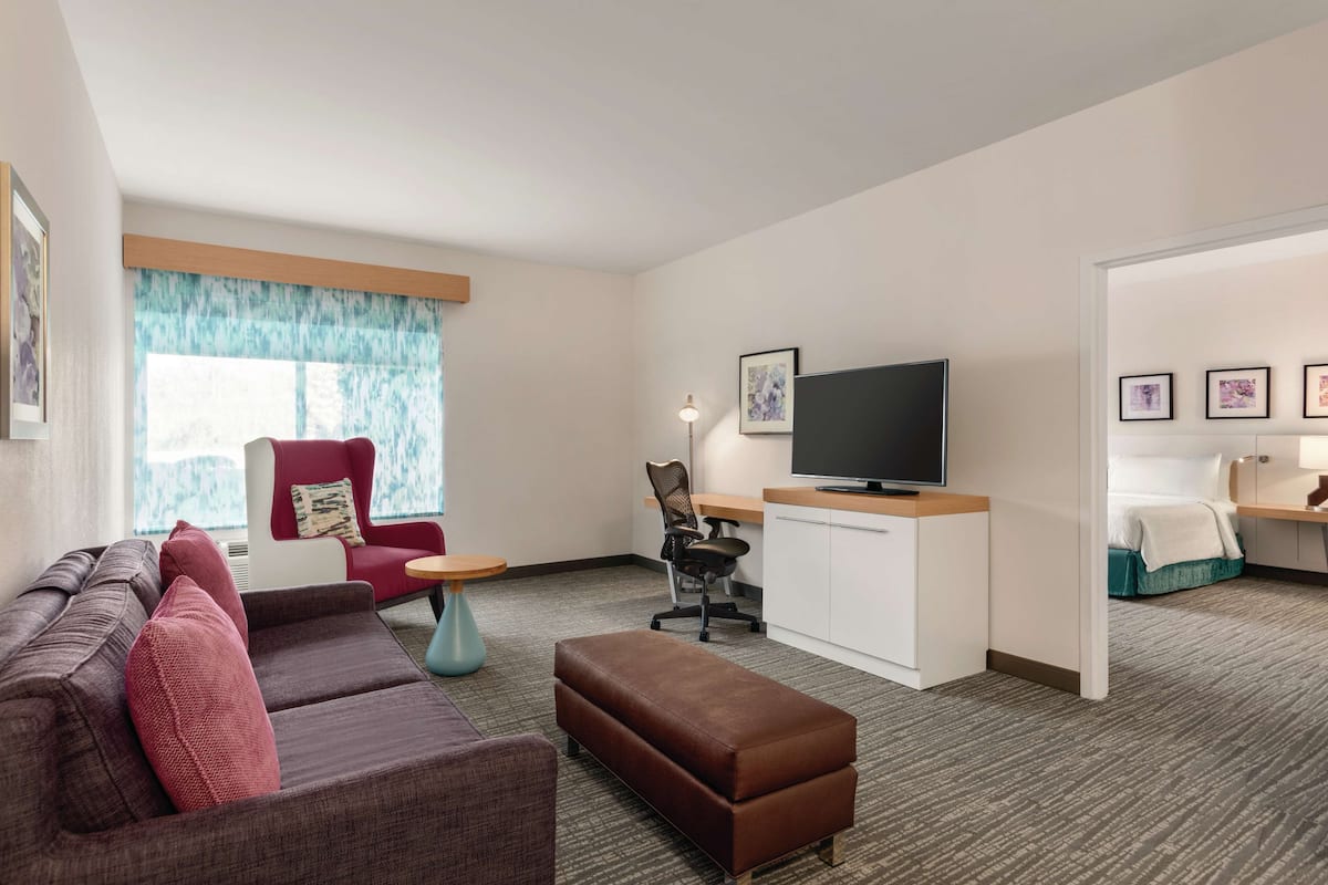 A modern hotel room with a sofa, ottoman, red armchair, desk, television, and artwork on the walls.