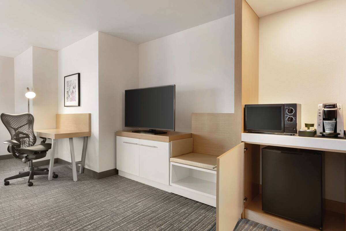 A hotel room with a desk, chair, television, microwave, and refrigerator.