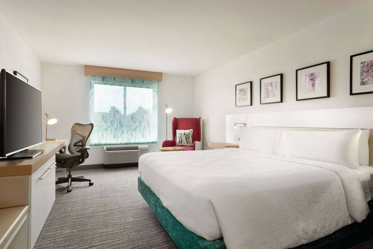 A hotel room with a bed, desk, chair, TV, and artwork on the wall.