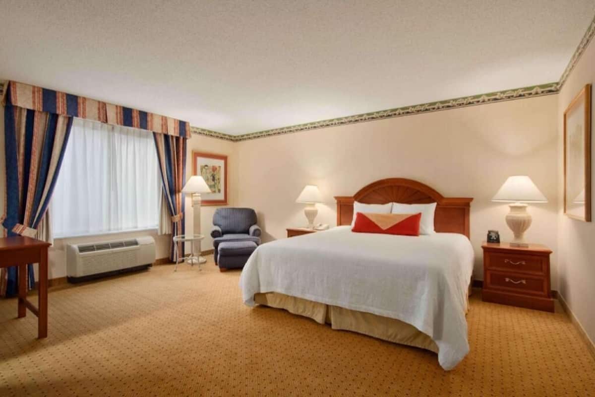 A hotel room with a bed, bedside lamps, a chair, a desk, and a window with curtains.