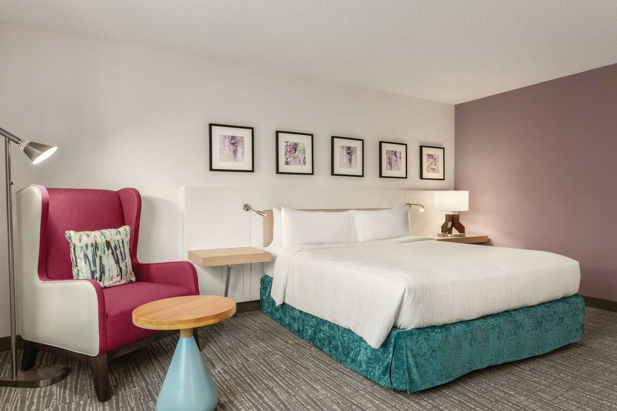 A hotel room with a bed, a chair, a small table, a lamp, and four framed pictures on the wall.