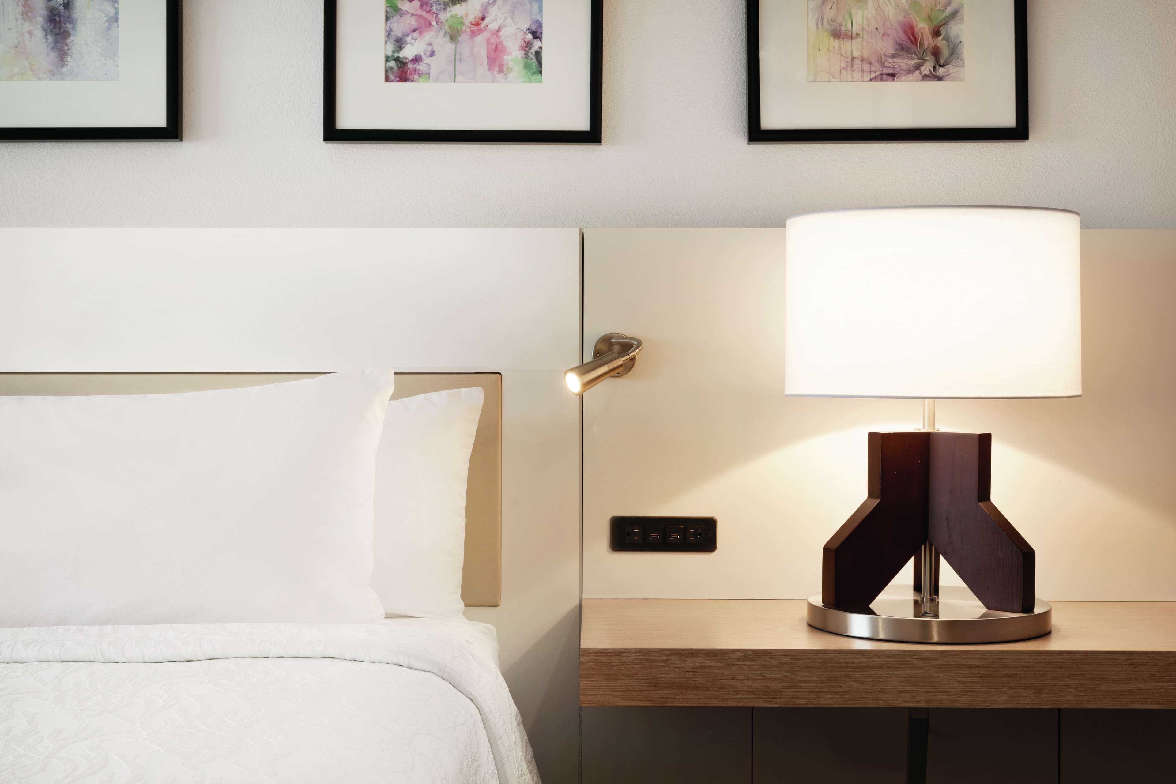 Un letto ben rifatto con un copriletto bianco e cuscini, un comodino con una lampada moderna e tre stampe floreali incorniciate a parete.