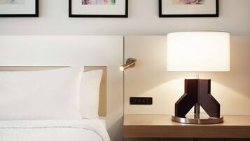 Un letto ben rifatto con un copriletto bianco e cuscini, un comodino con una lampada moderna e tre stampe floreali incorniciate a parete.