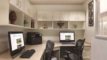 A home office with two desktop computers, a printer, and a bookshelf with decorative items.
