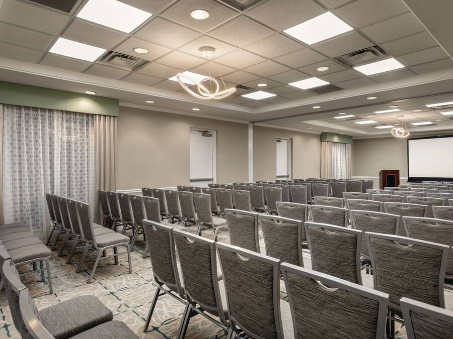 Un salón de conferencias con filas de sillas, una pantalla de proyector y un amplio ventanal con cortinas.