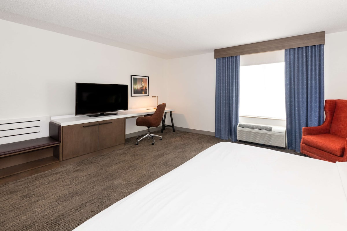 A hotel room with a bed, a desk with a TV, a chair, a red armchair, and a window with curtains.