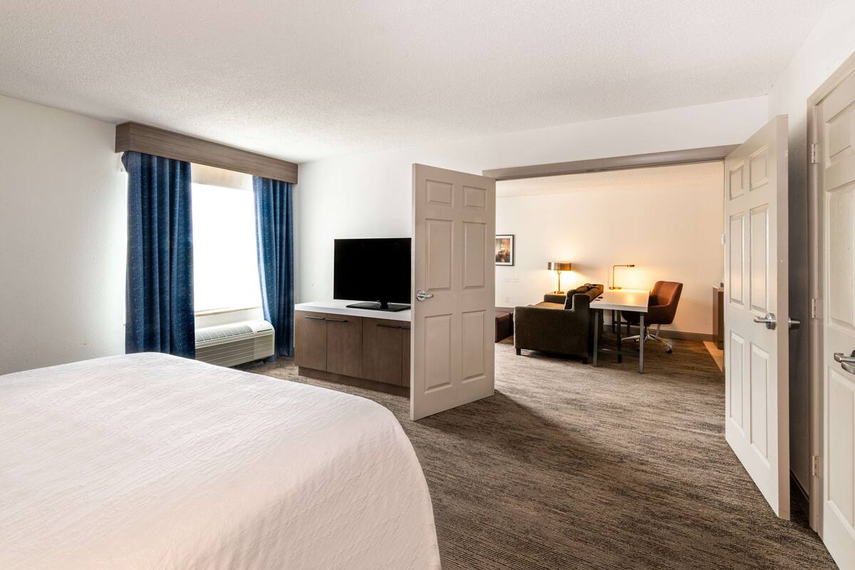 A hotel room with a bed, a desk with a computer, a television, and a door leading to another room.