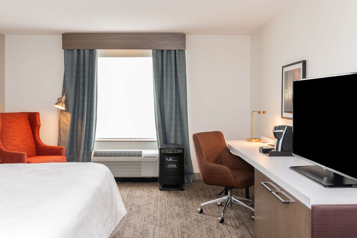 A hotel room with a bed, two chairs, a desk with a computer, a TV, and a window with curtains.