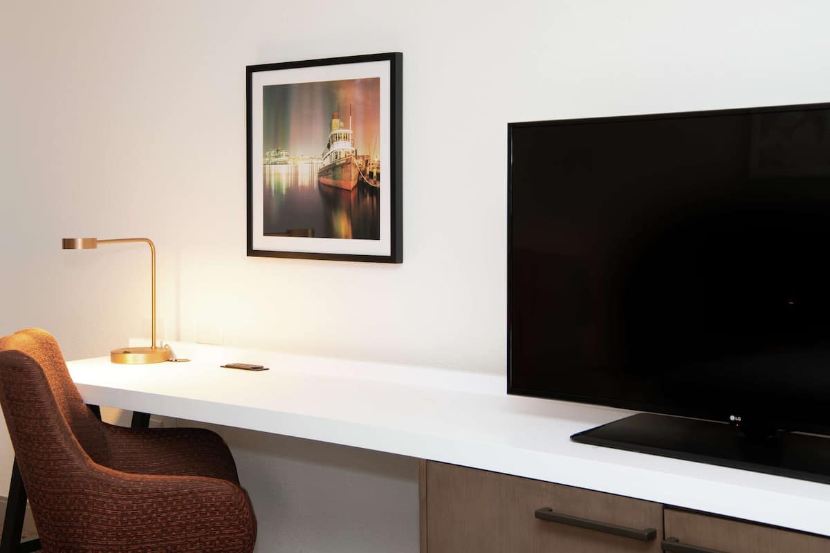 A modern hotel room with a white desk, a large flat-screen TV, a framed picture of a cityscape, a brown upholstered chair, and a desk lamp.