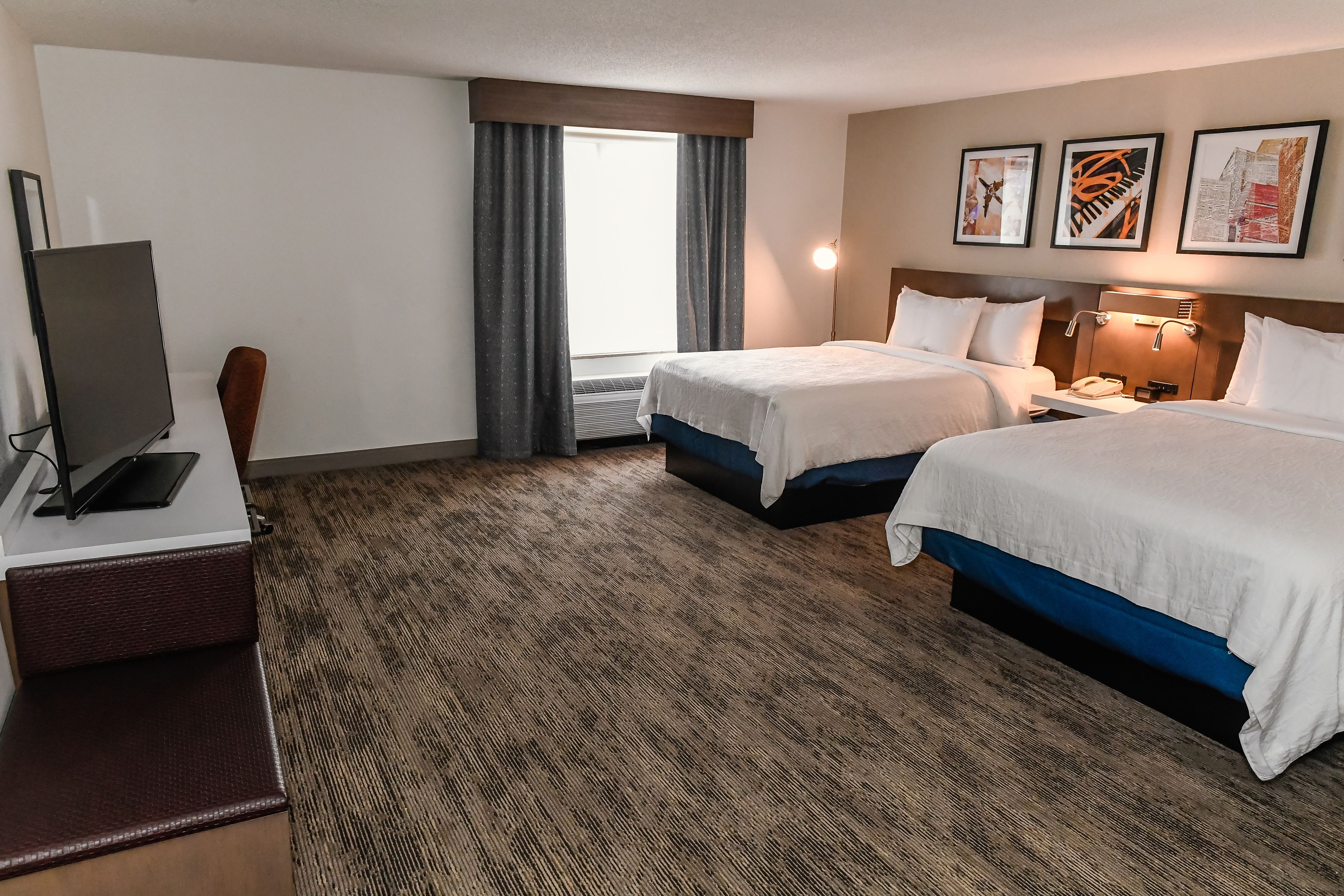 A hotel room with two beds, a desk with a computer, a chair, a window with curtains, and artwork on the wall.
