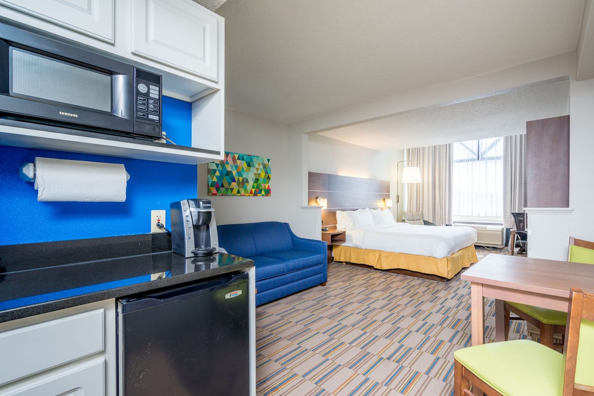 A hotel room with a kitchenette, a bed, a blue sofa, a wooden table, and a colorful abstract painting on the wall.
