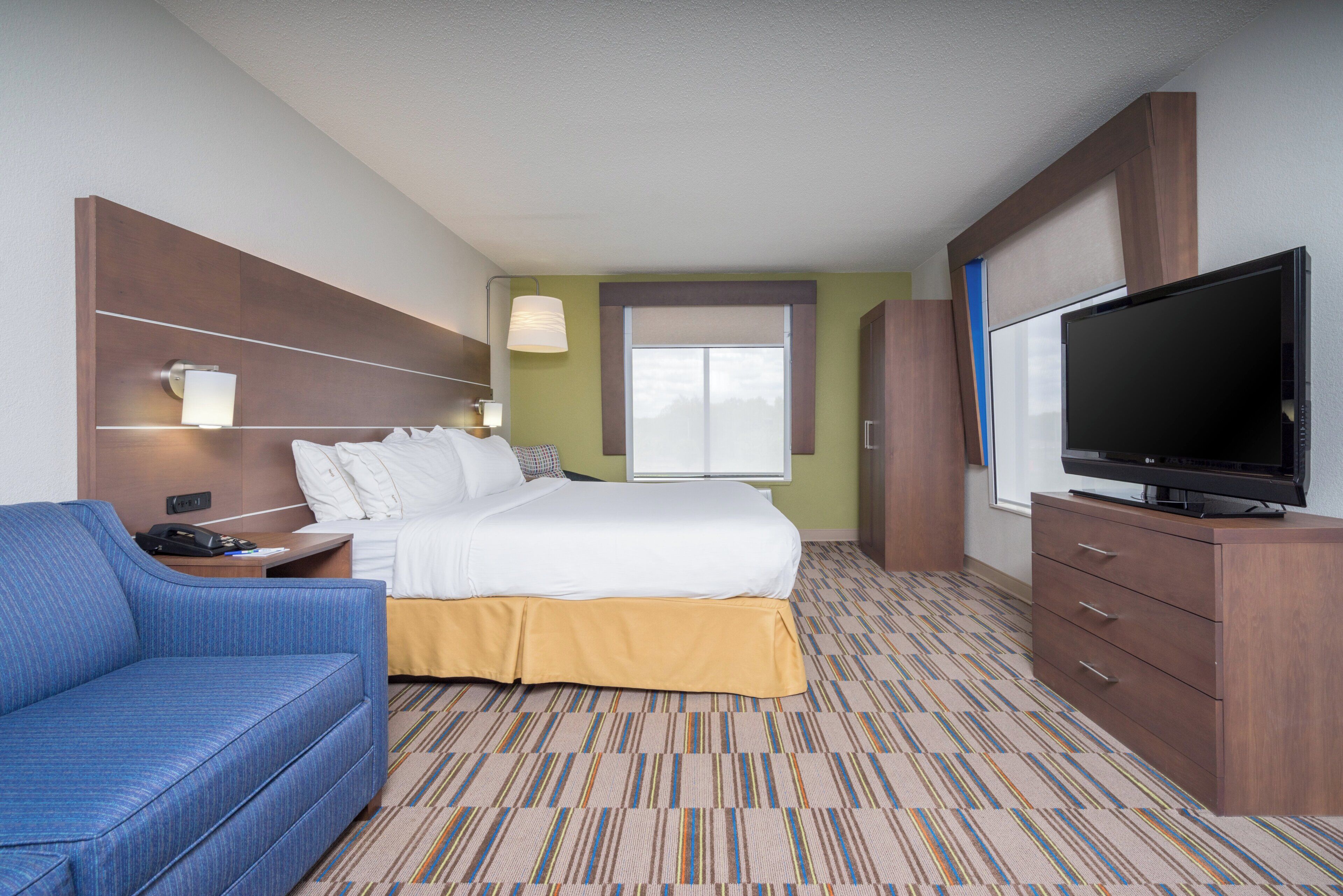 Une chambre d’hôtel avec un lit, une télévision, un fauteuil bleu, un bureau avec une lampe et une fenêtre avec des stores.