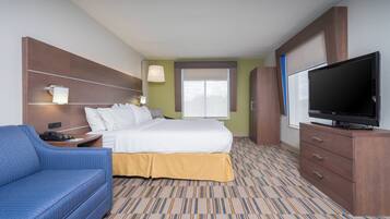 Une chambre d’hôtel avec un lit, une télévision, un fauteuil bleu, un bureau avec une lampe et une fenêtre avec des stores.