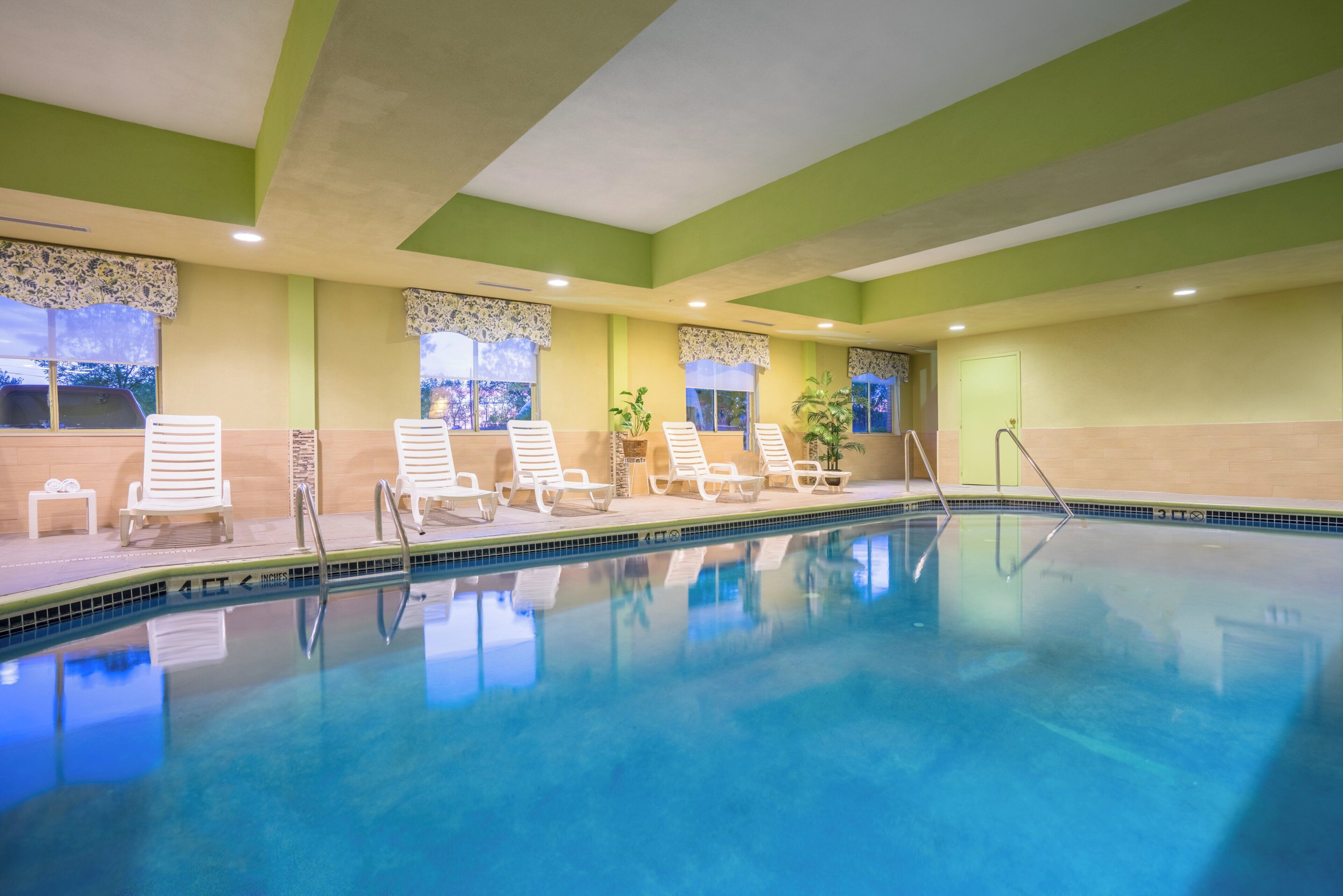 Una piscina interior con agua cristalina y azul, rodeada de sillas reclinables blancas y un suelo de baldosas.
