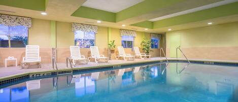 Una piscina interior con agua cristalina y azul, rodeada de sillas reclinables blancas y un suelo de baldosas.