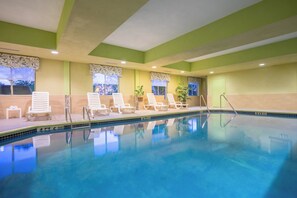An indoor swimming pool with clear blue water, surrounded by white lounge chairs and a tiled floor.