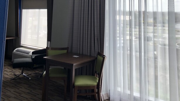 Una camera d'albergo con un tavolo da pranzo, sedie, una ventola a soffitto e un'ampia finestra con tende leggere.