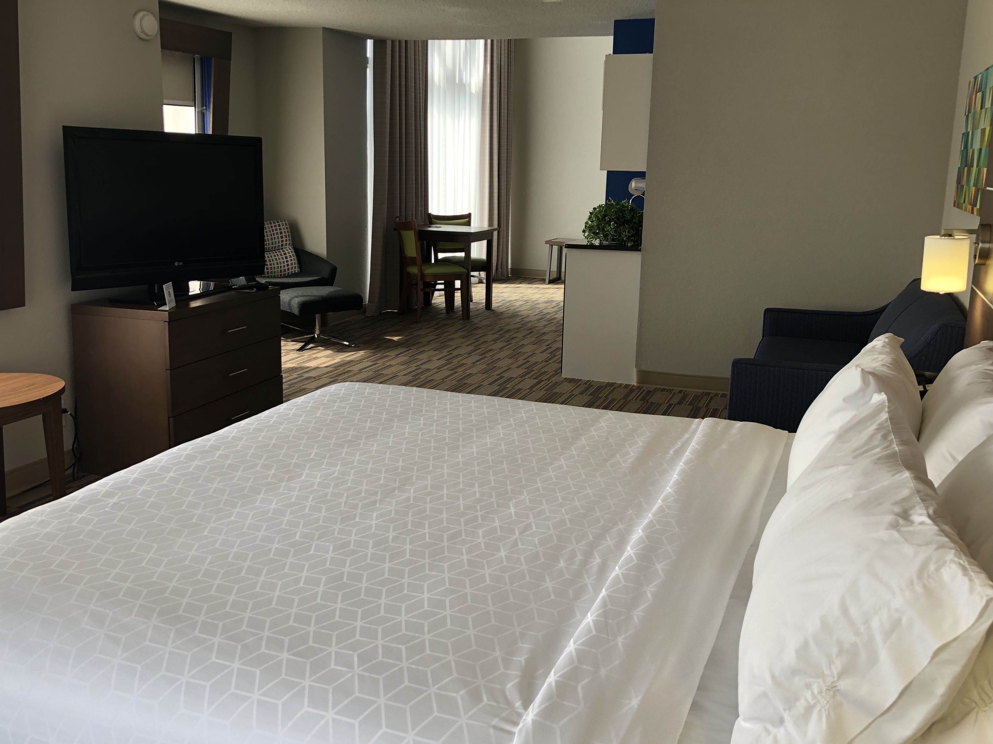 A hotel room with a large bed, a desk with a TV, a small dining area, and a seating area with a plant.