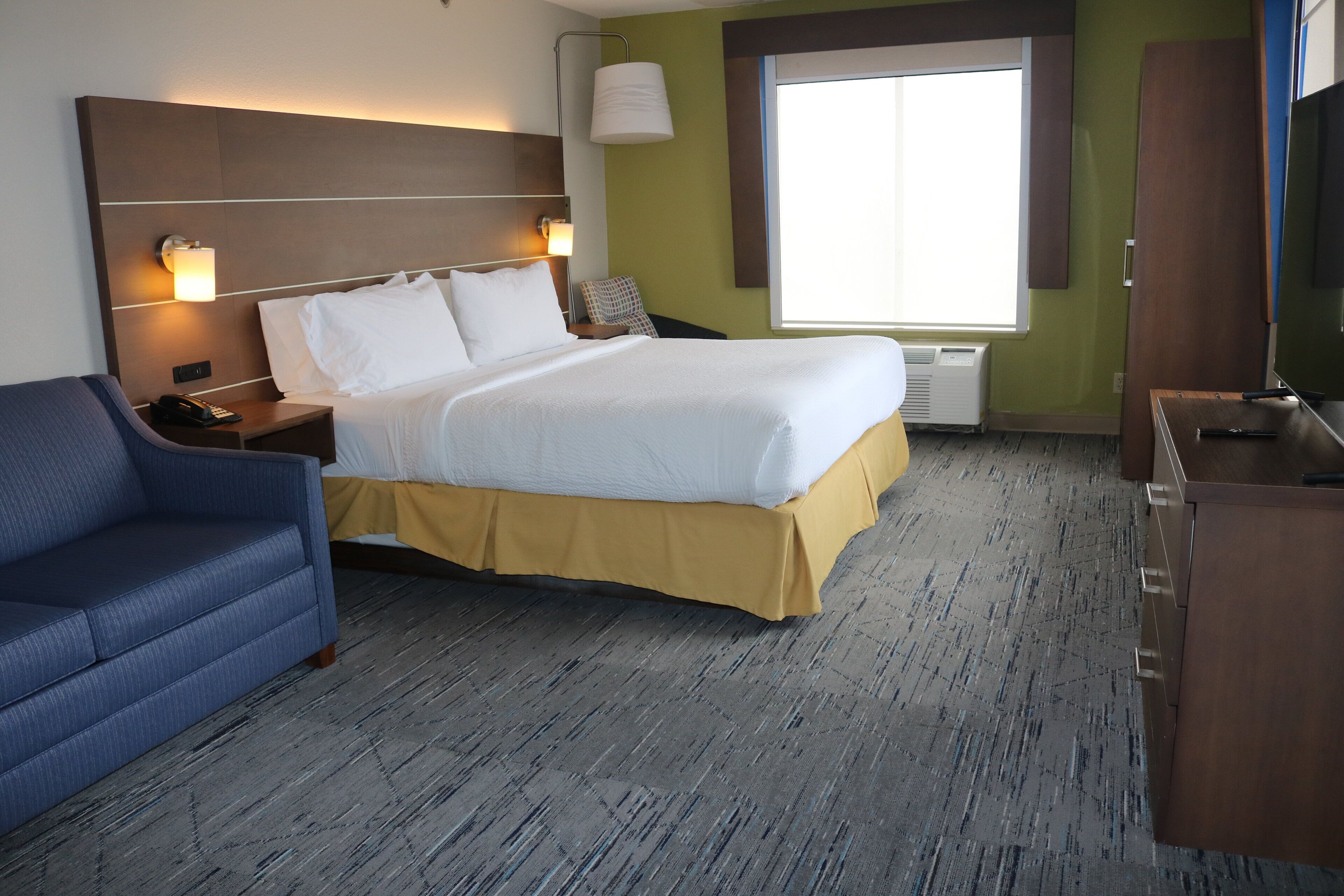 Een hotelkamer met een bed, een blauwe fauteuil, een bureau met een televisie en een raam met zonwering.