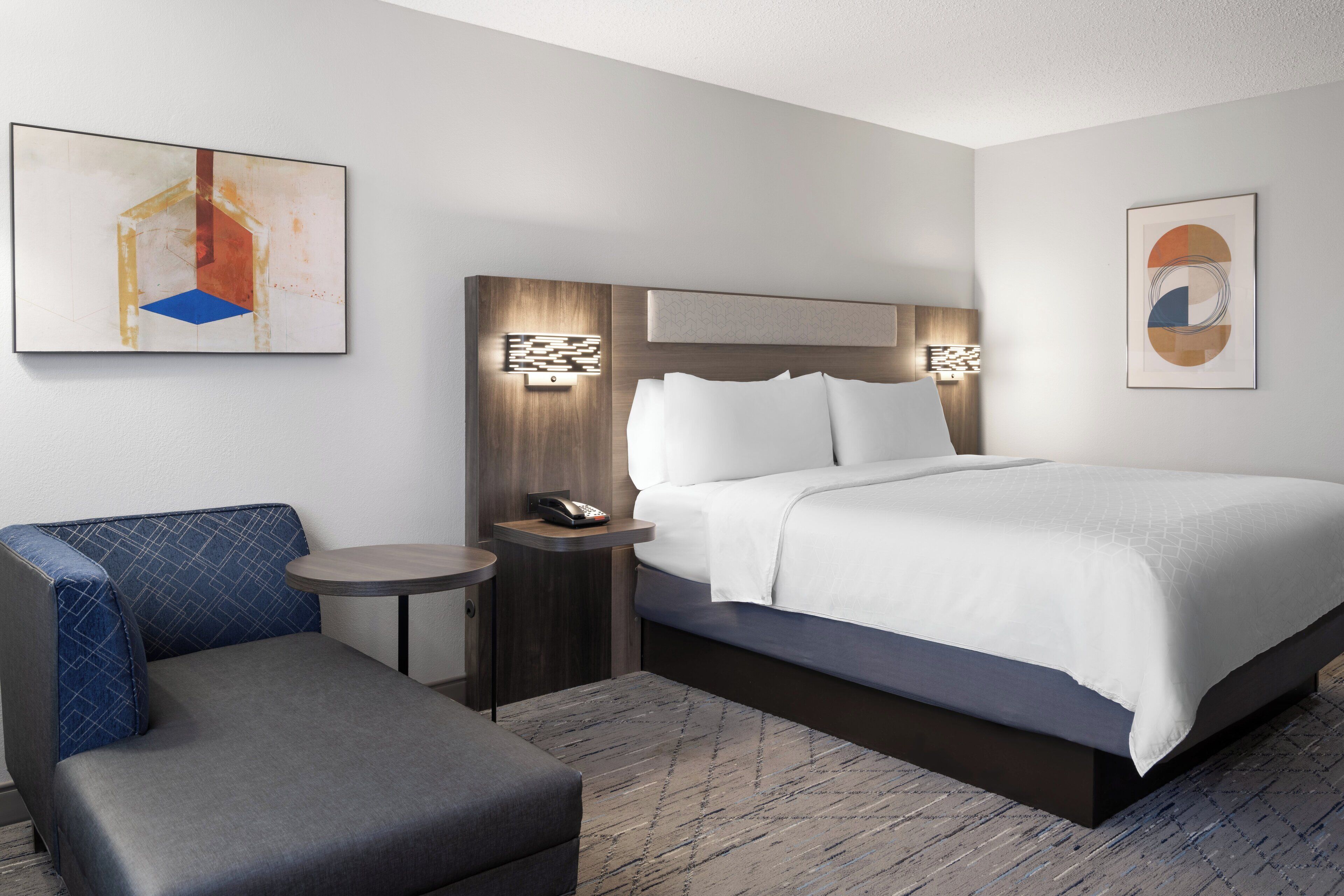 A hotel room with a bed, a seating area, a bedside table with a phone, and artwork on the walls.