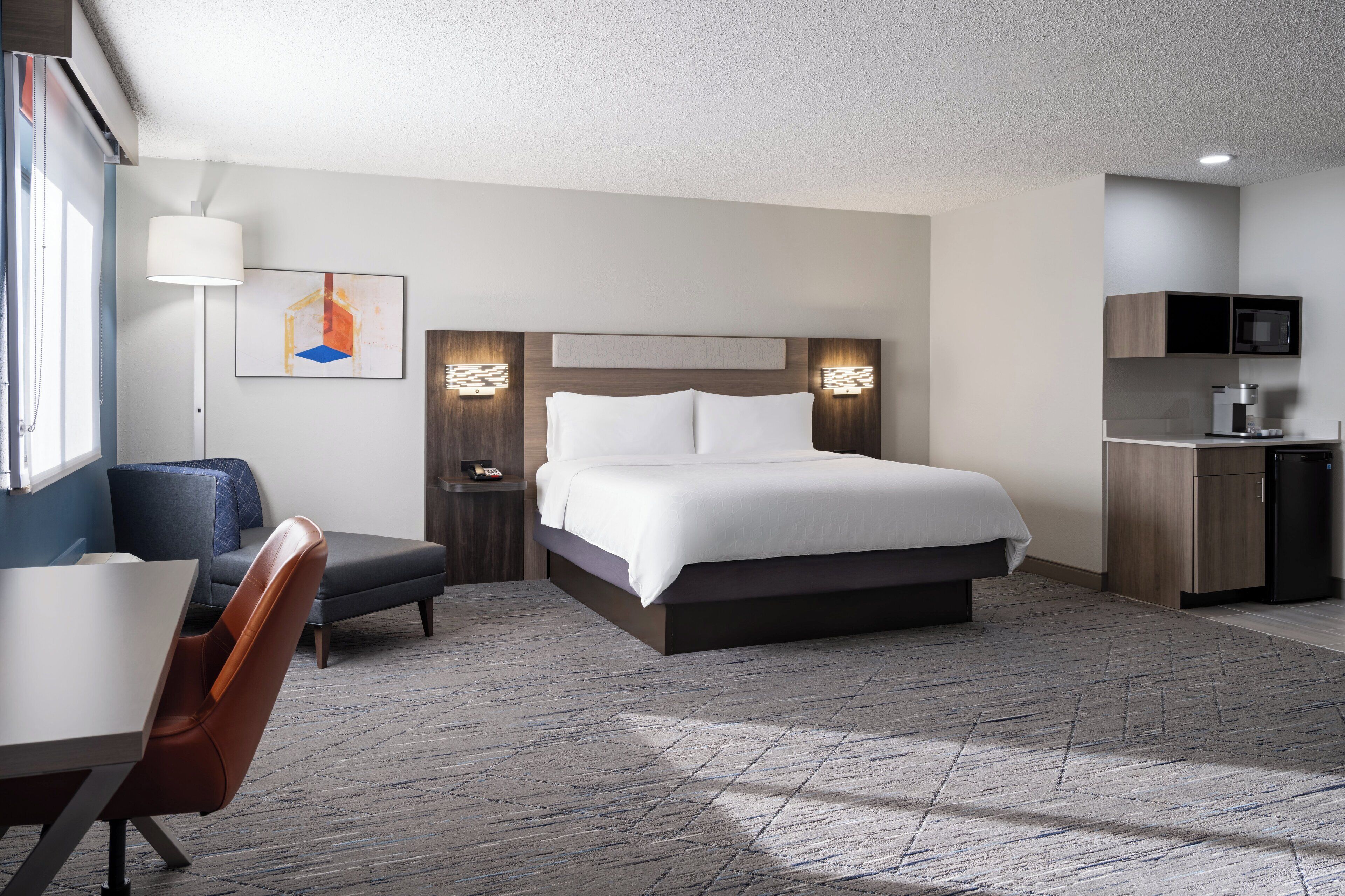 A modern hotel room with a large bed, a desk, a chair, a small kitchenette, and a painting on the wall.