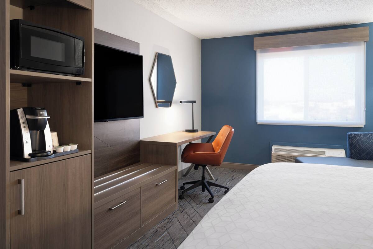A hotel room with a bed, a desk, a chair, a microwave, a coffee maker, a TV, and a window with blinds.