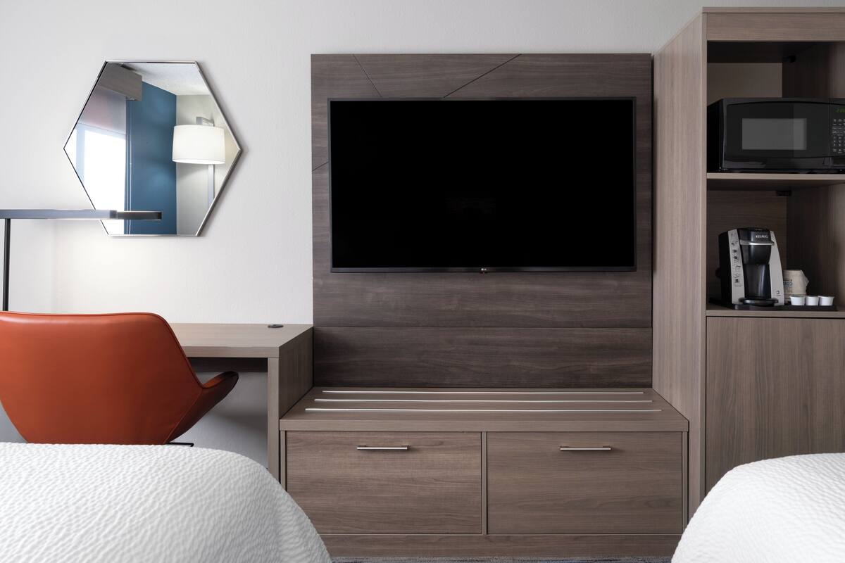 A hotel room with a large flat-screen TV mounted on a wooden cabinet, a desk with a chair, and a microwave on a shelf.