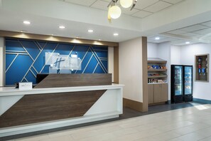 A reception area with a modern design, including a reception desk, a blue and white wall with geometric patterns, and a refrigerator stocked with beverages. - Holiday Inn Express & Suites Midwest by IHG (Midwest City)