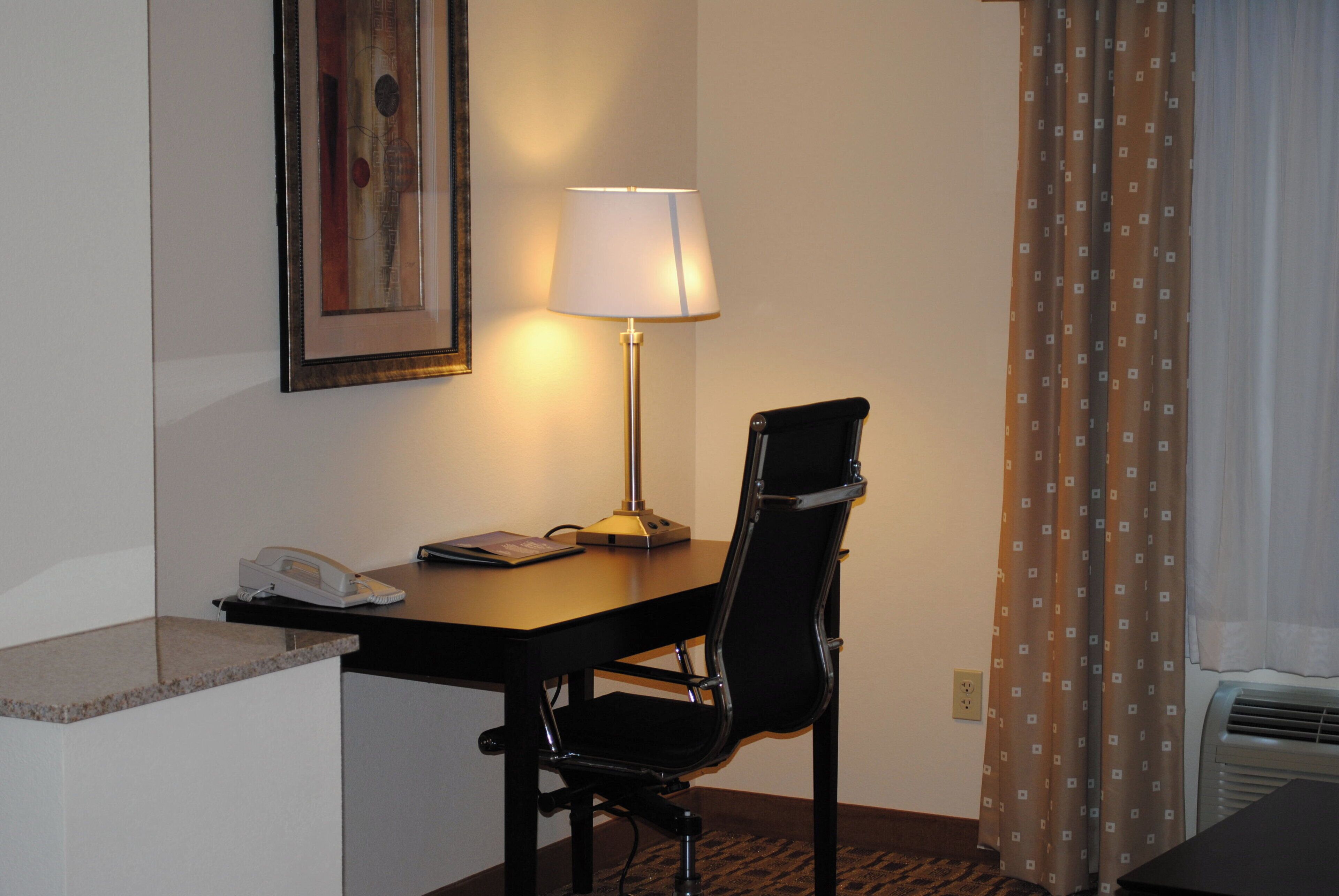 Ett hotellrum med ett skrivbord, en stol, en lampa, en telefon och en tavla på väggen.