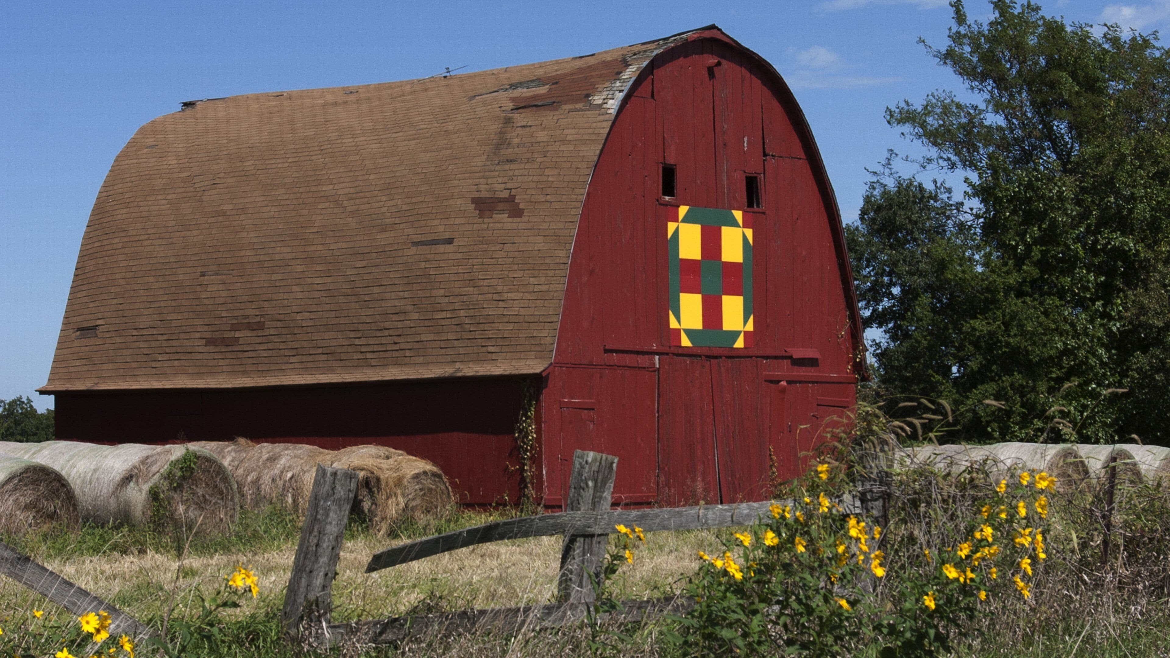 A red barn with a distinctive window design, surrounded by hay bales and wildflowers.