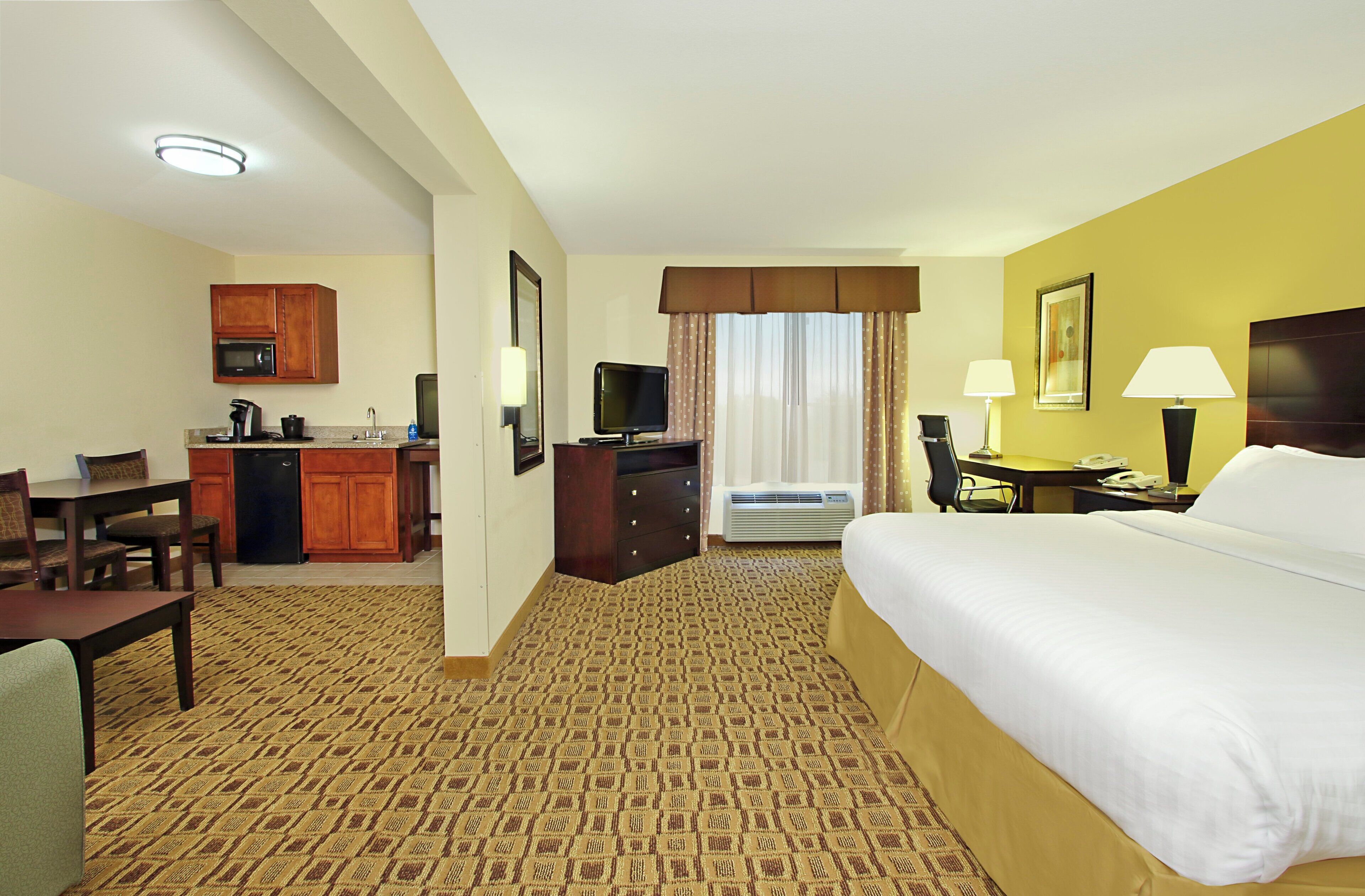 A hotel room with a bed, desk, chair, kitchenette, and a television.