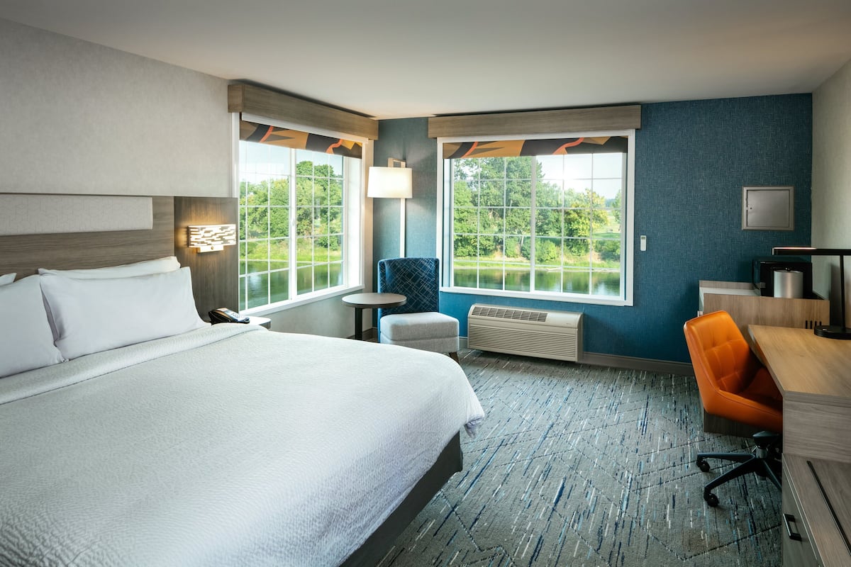 Une chambre d’hôtel avec un grand lit, un bureau, une chaise orange, une petite table et deux fenêtres donnant sur un paysage verdoyant.