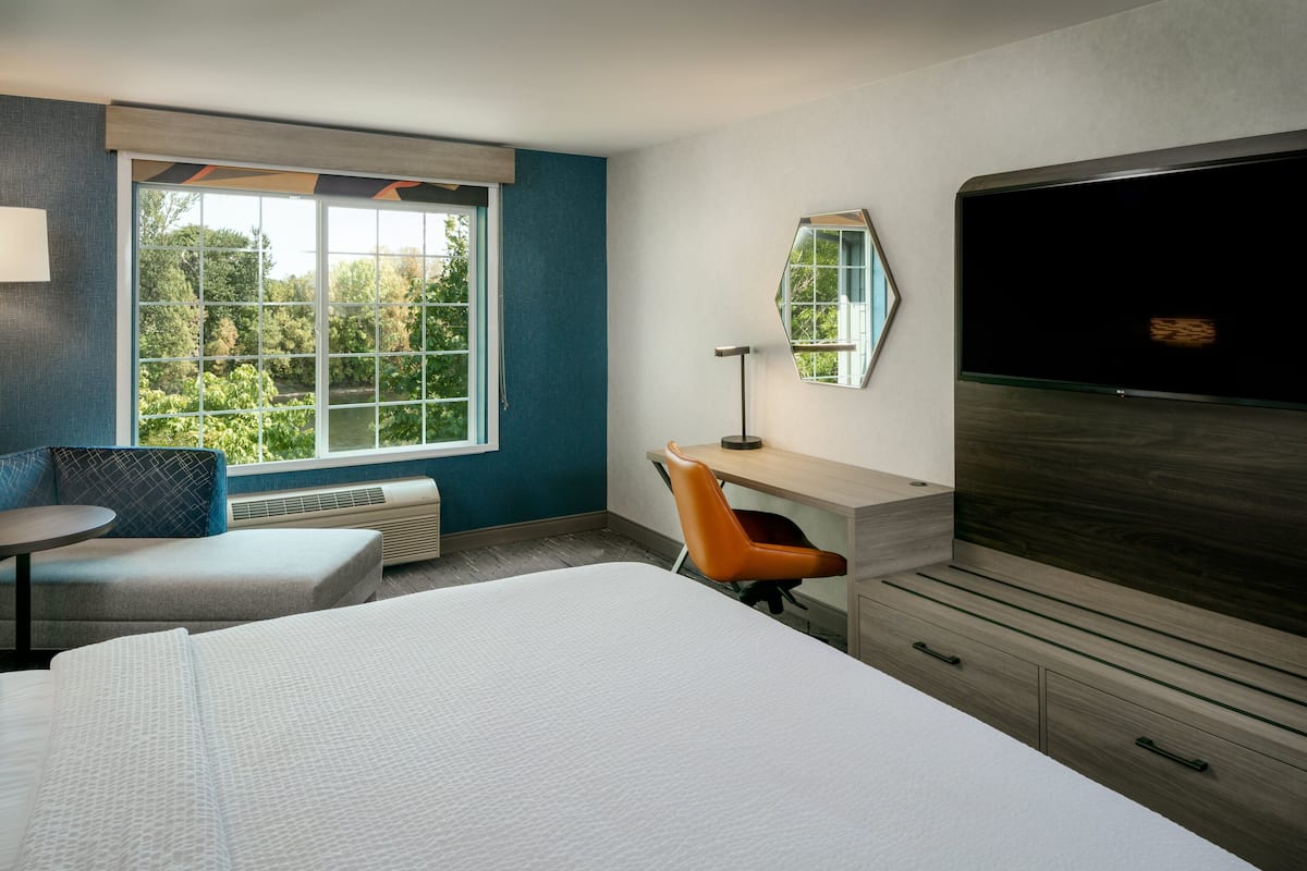 Une chambre d’hôtel avec un grand lit, un bureau, une chaise, une télévision et une fenêtre donnant sur des arbres.