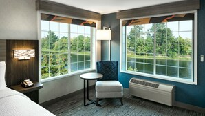 A hotel room with a large window offering a view of a lake and trees, a small round table, a blue chair, a bed, and a wall-mounted phone. - Holiday Inn Express Corvallis-On the River by IHG (Corvallis)