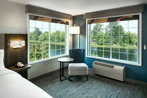 A hotel room with a large window offering a view of a lake and trees, a small round table, a blue chair, a bed, and a wall-mounted phone. - Holiday Inn Express Corvallis-On the River by IHG (Corvallis)