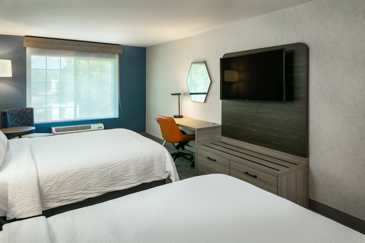 Une chambre d’hôtel avec deux lits, un bureau, une chaise, un téléviseur à écran plat et une fenêtre avec des stores.