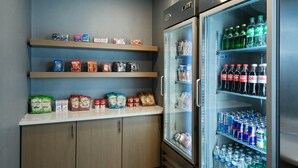 Una kitchenette con estantes llenos de bocadillos y un refrigerador que contiene agua embotellada y refrescos.