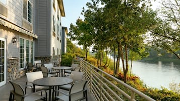 Un patio con mobiliario exterior que da a un lago, con una pared de piedra y un edificio de amplios ventanales.