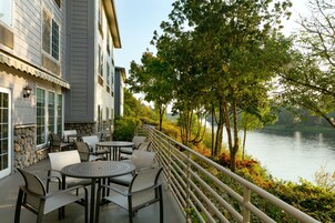 Un patio con mobiliario exterior que da a un lago, con una pared de piedra y un edificio de amplios ventanales.