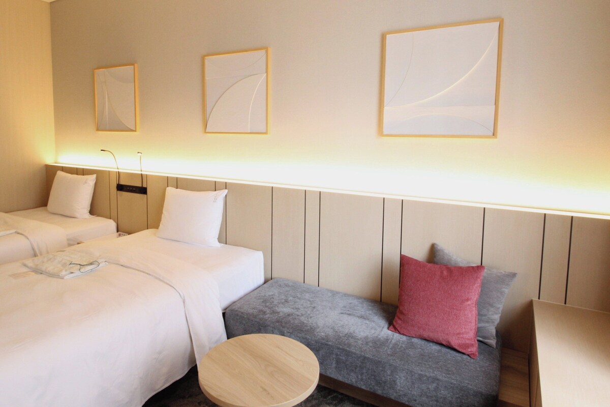 A hotel room with two beds, a grey sofa with a red pillow, a round wooden table, and wall-mounted artwork.