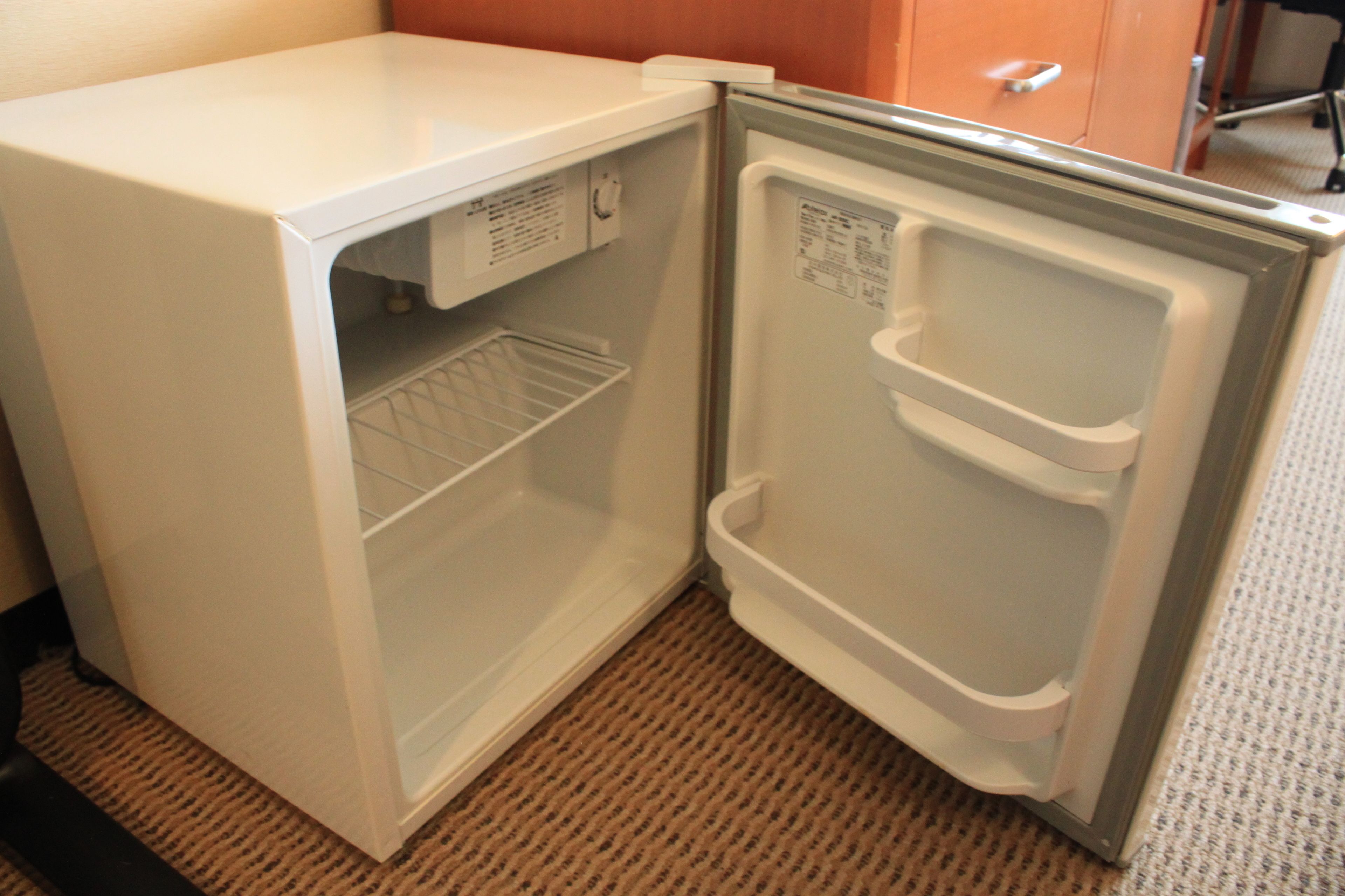 A compact white refrigerator with a freezer compartment, located in a room with a patterned carpet and a wooden cabinet in the background.